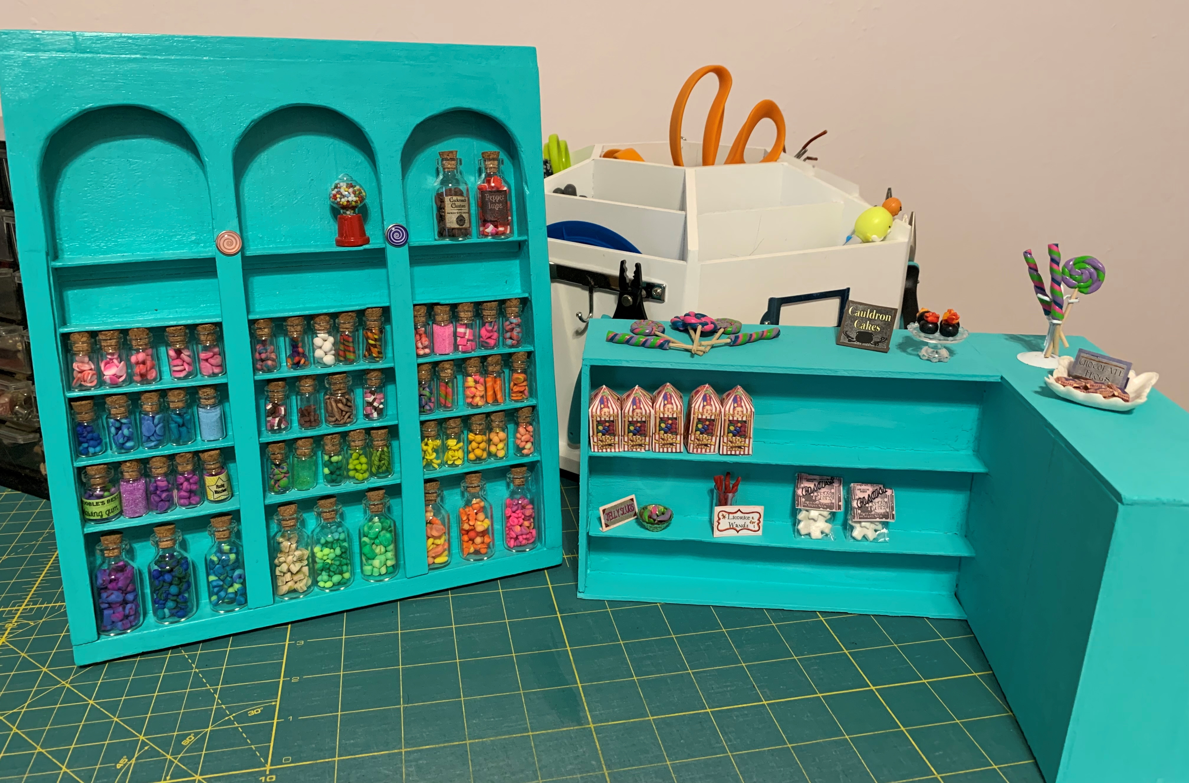 A turquoise floor to ceiling shelf and counter for a miniature candy shop. The shelves are partly filled with jars and packs of candy. 