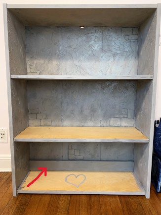 A bookcase with the interior back and sides painted to look like gray castle walls. 