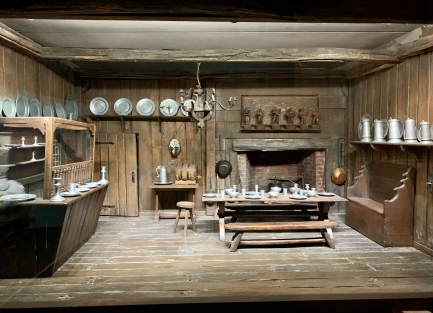 A dollhouse kitchen with wooden furniture and pewter dishes.